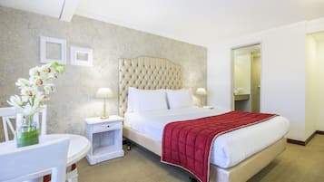 Una habitación de hotel con cabecera capitonada, una cama grande con manta roja, una mesita de noche con lámpara, una pequeña mesa con un jarrón de flores y una silla.