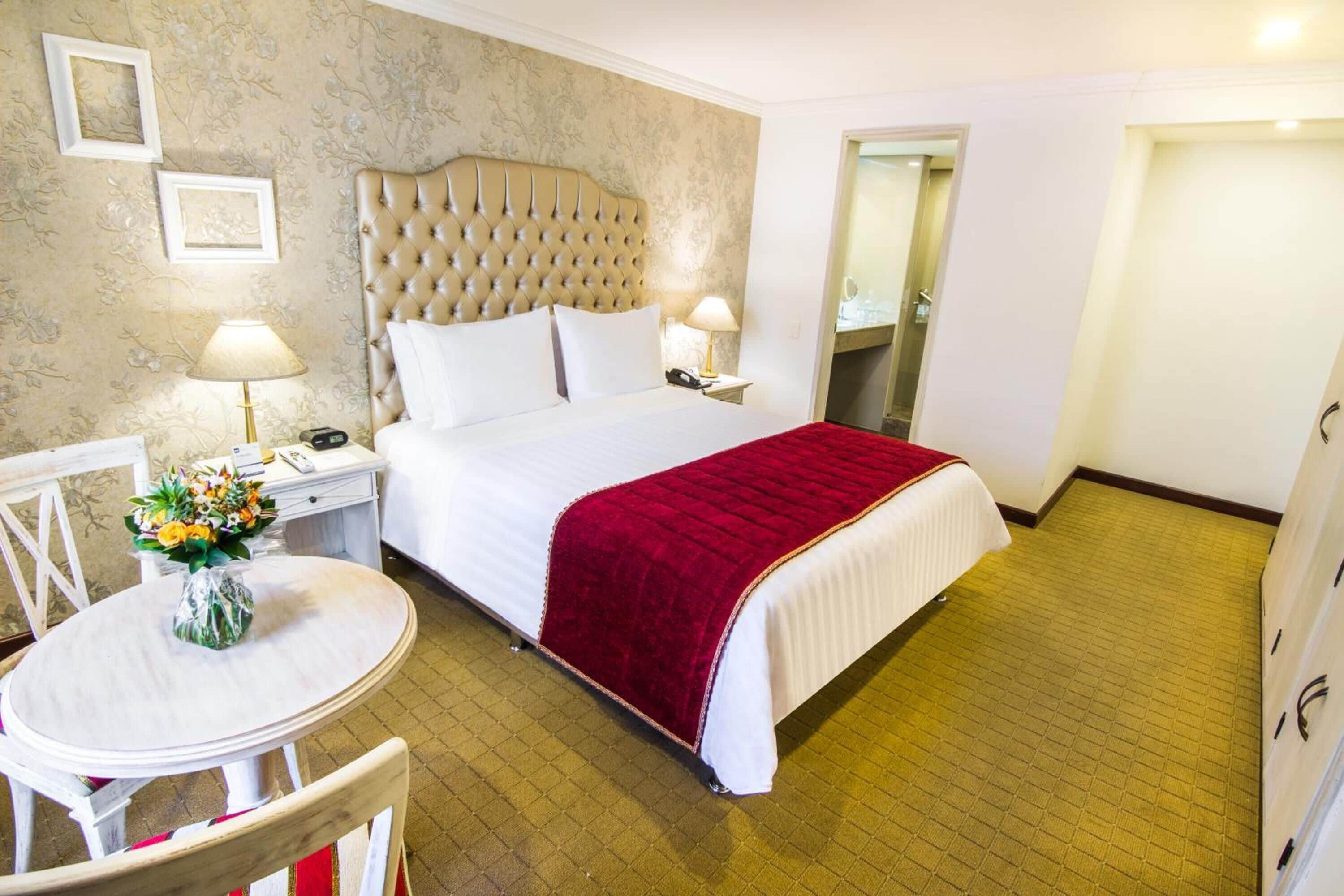 A hotel room with a large bed, a bedside table with a lamp, a chair, and a small table with a vase of flowers.