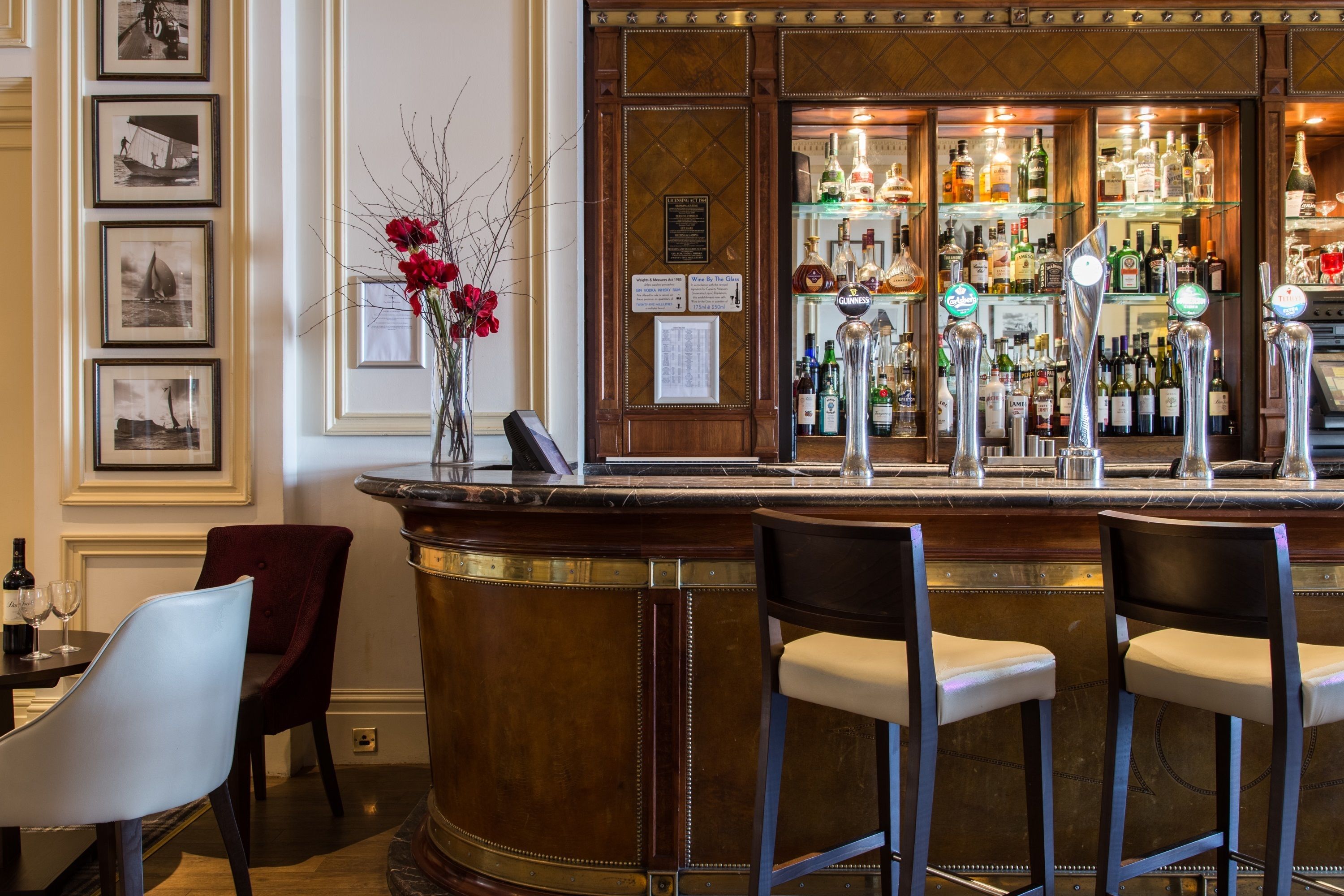 A bar area with a marble counter, two bar stools, a vase with red flowers, and a wall with framed pictures.