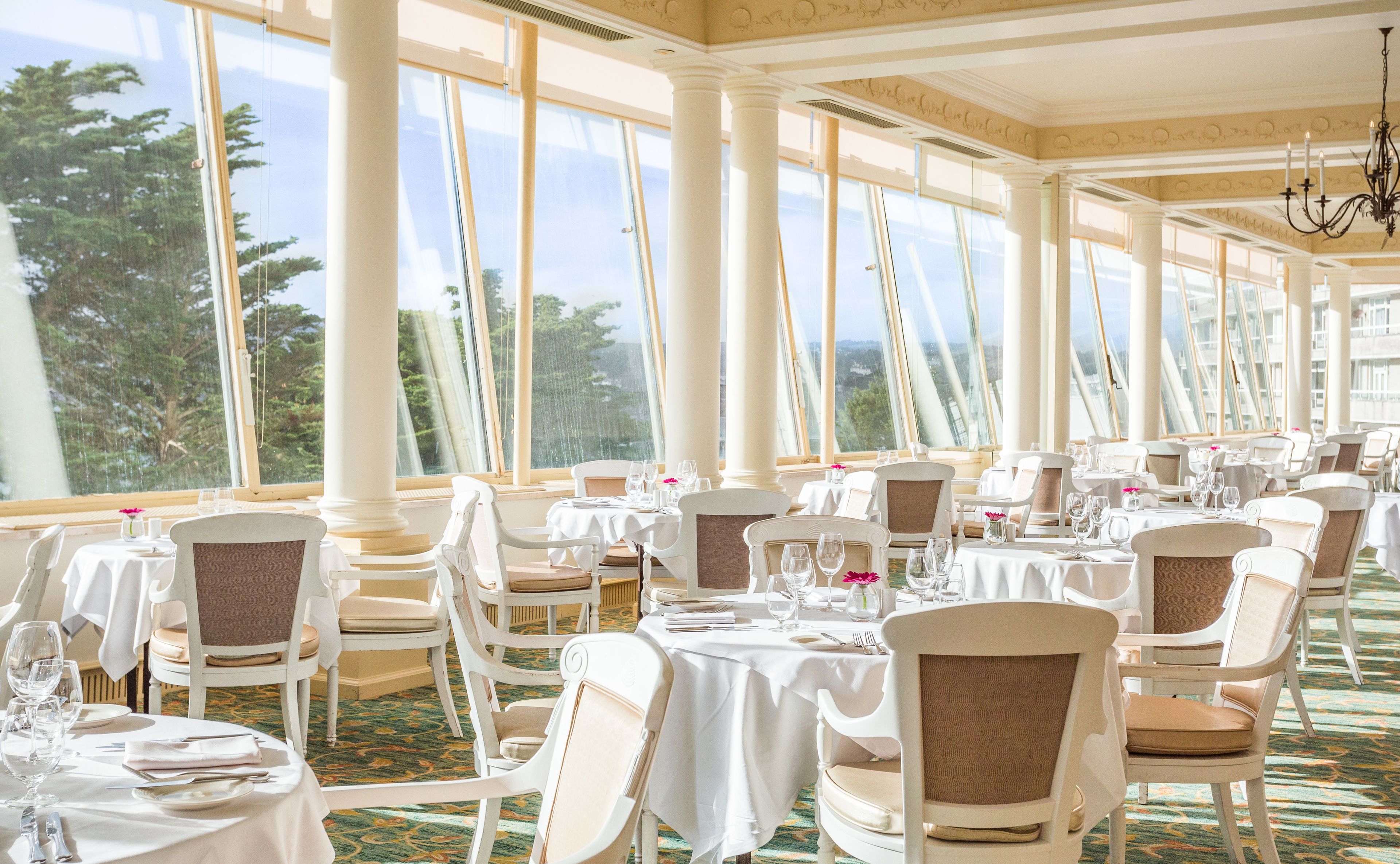 Un restaurant avec des tables et des chaises blanches, de grandes fenêtres et un lustre.