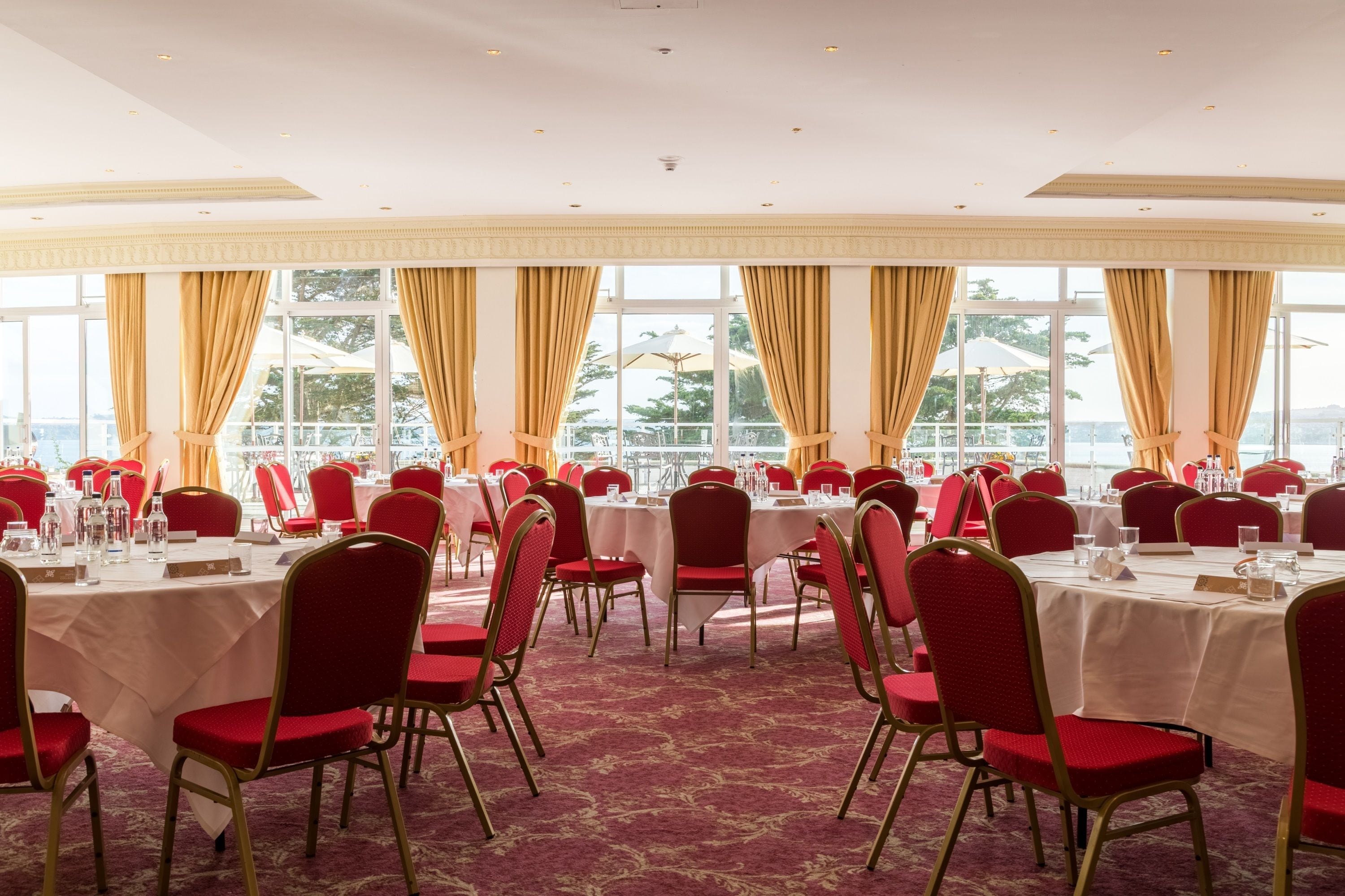 Un salón de banquetes con mesas redondas y sillas rojas, ventanas amplias con cortinas y vistas al exterior.