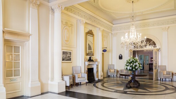 A grand lobby with classical architecture, including columns, a chandelier, and a fireplace.