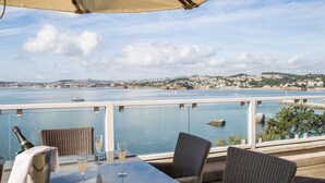 A terrace with wicker furniture, a table with a bottle and glasses, and a view of a coastal town across the water.