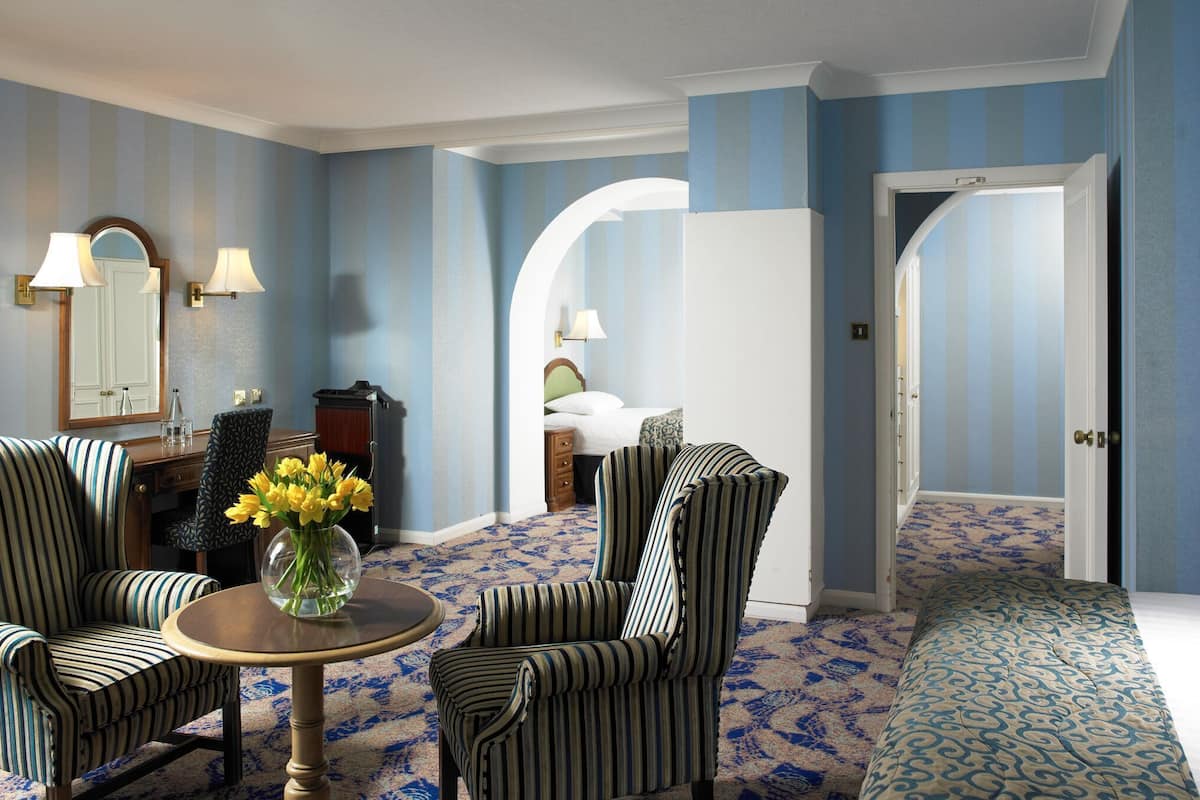 Una habitación con sillas a rayas, una mesa redonda con un jarrón de flores, una cama con colcha estampada y una puerta que da a otra habitación.