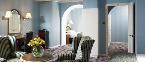 A room with striped chairs, a round table with a vase of flowers, a bed with a patterned bedspread, and a door leading to another room.