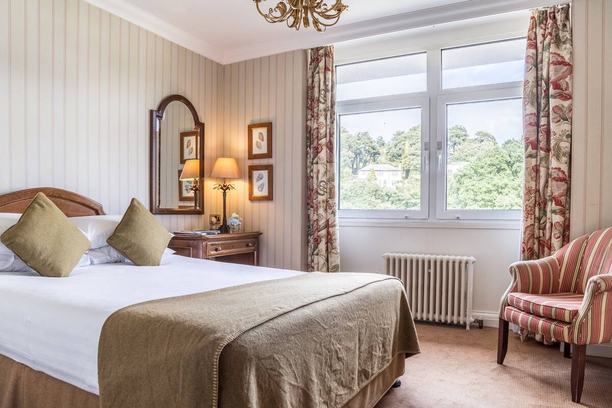 Una habitación de hotel con una cama grande, una silla, un escritorio con lámpara, un espejo y una ventana con cortinas.