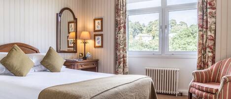 Une chambre d’hôtel avec un grand lit, une chaise, un bureau avec une lampe, un miroir et une fenêtre avec des rideaux.