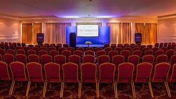 A conference room with rows of red chairs, a stage with a screen displaying 'The Imperial Torquay', and two speakers on either side.
