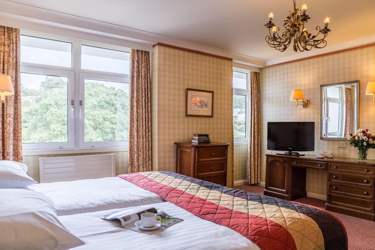 Una habitación de hotel con una cama grande, un escritorio con televisor, un aparador y una ventana con cortinas.