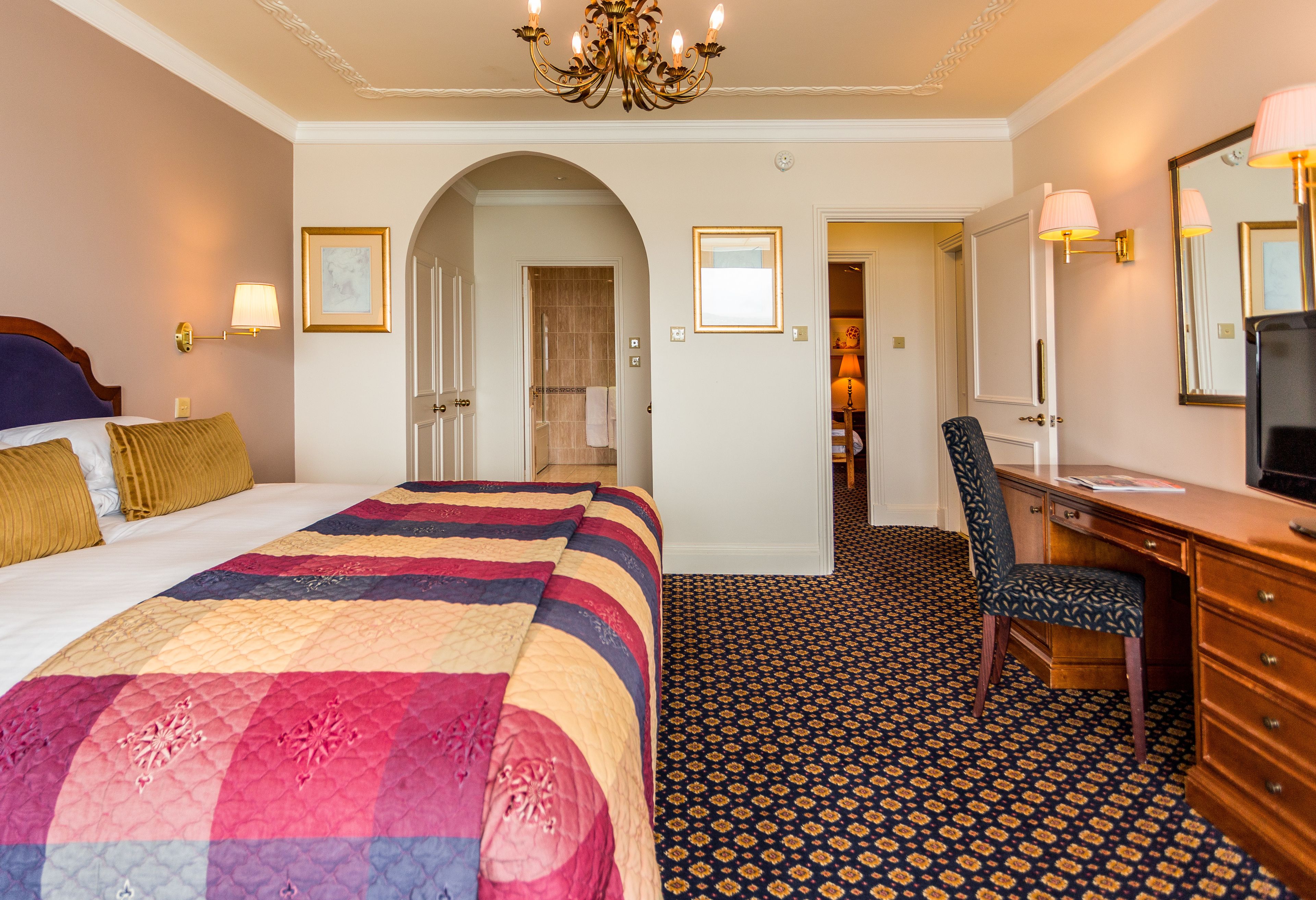 A hotel room with a large bed, a desk, a chair, a mirror, and a chandelier.