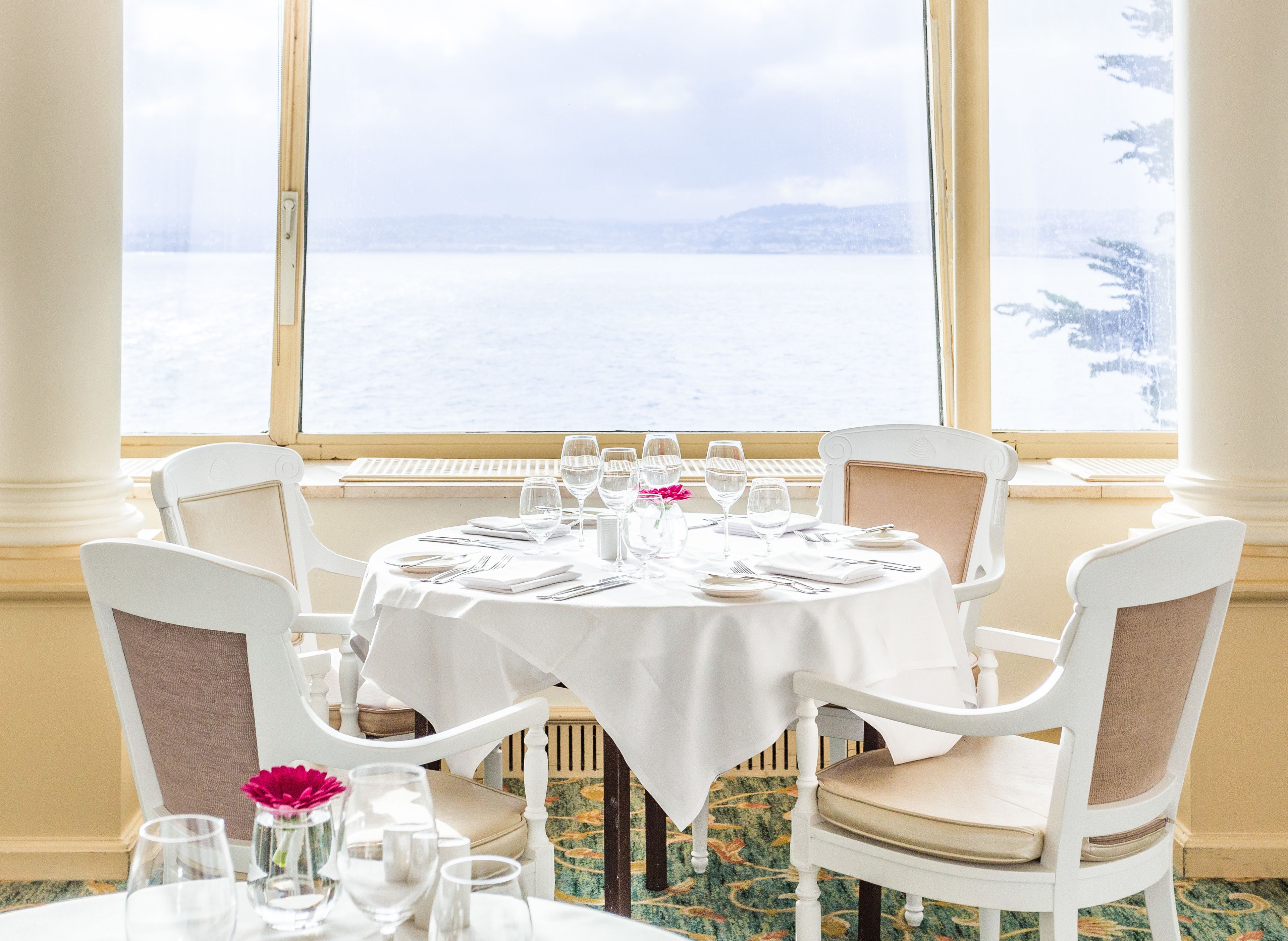 Mesa de comedor para cuatro con manteles blancos, sillas y cristalería, con vista a un paisaje nevado a través de amplios ventanales.