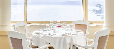 Mesa de comedor para cuatro con manteles blancos, sillas y cristalería, con vista a un paisaje nevado a través de amplios ventanales.