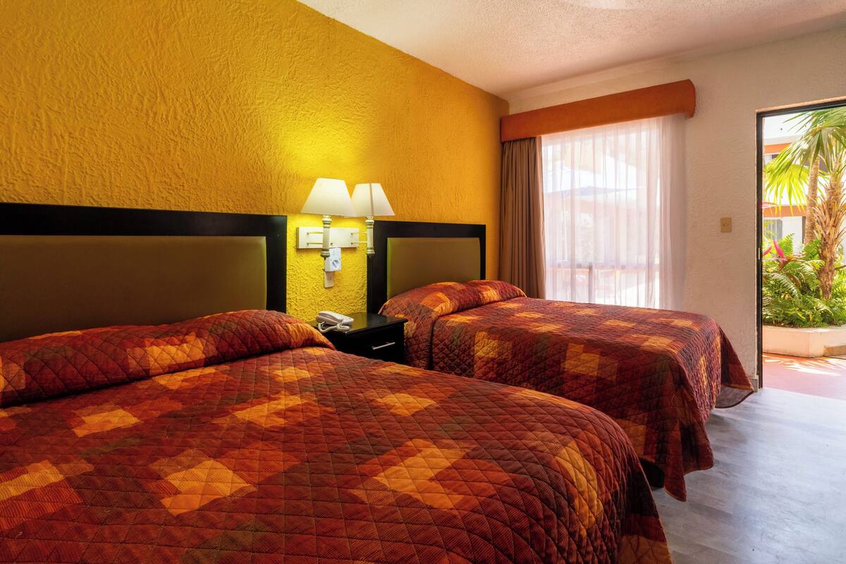A hotel room with two beds, a nightstand, a lamp, and a view of greenery outside.