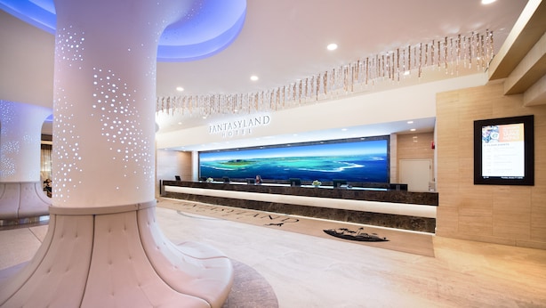 Un hall d’hôtel moderne, doté d’un grand comptoir d’accueil, d’un plafond haut orné d’un éclairage décoratif et offrant une vue sur l’océan grâce à une grande fenêtre.