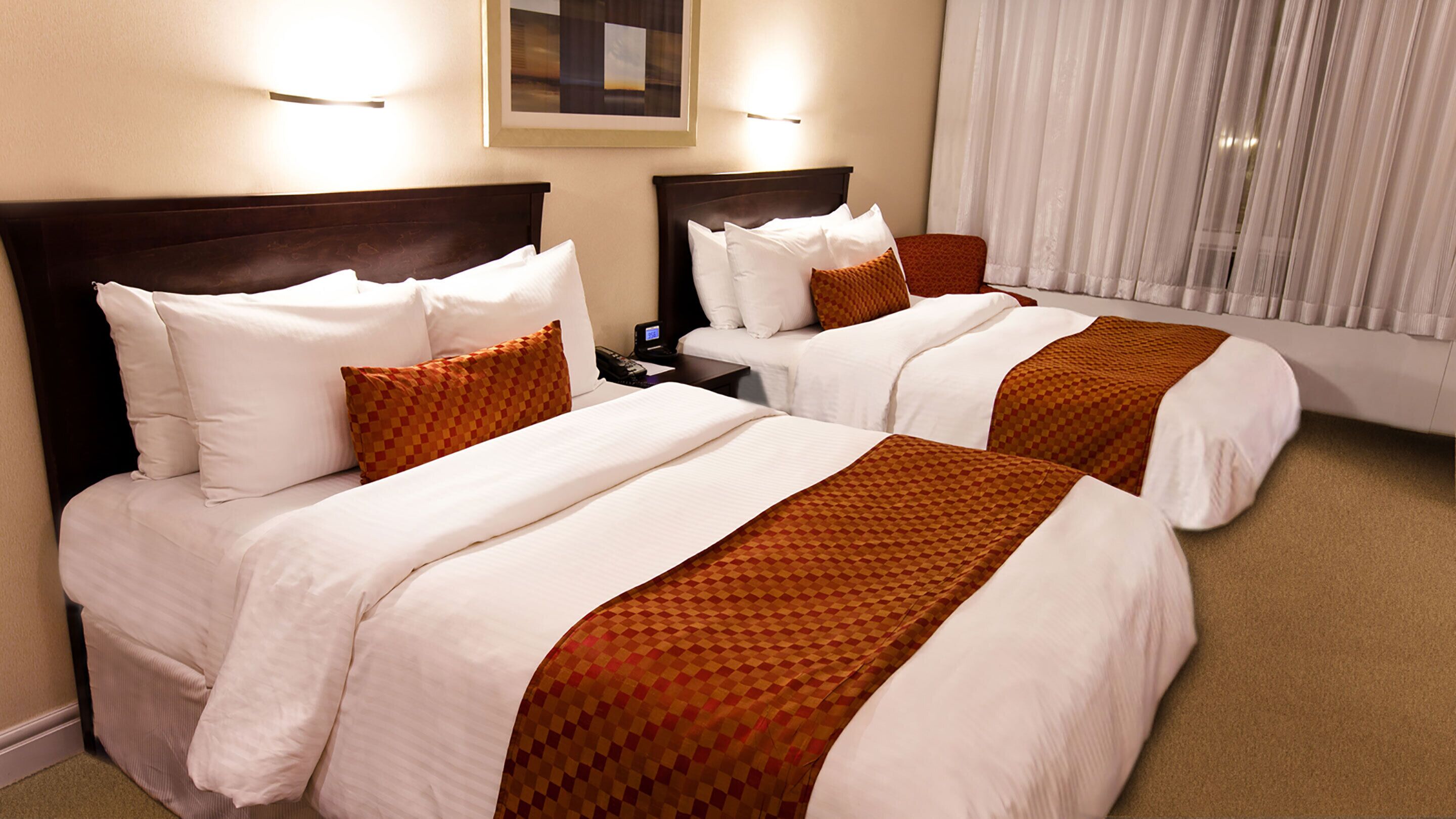 Una habitación de hotel con dos camas, cada una con sábanas blancas y mantas marrones a cuadros. Hay mesitas de noche con lámparas y un cuadro en la pared.