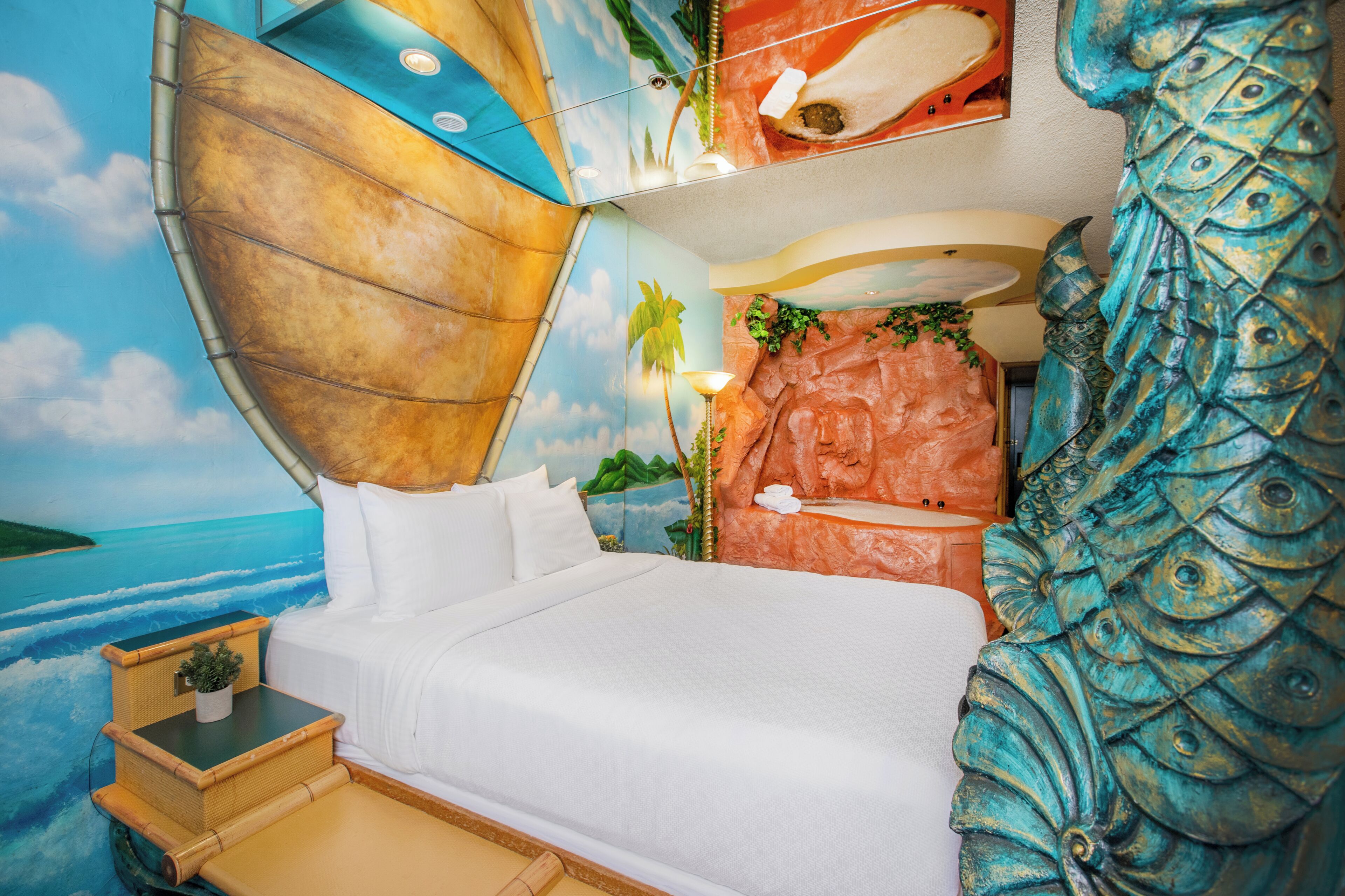 A room with a bed, a small table with a plant, a wall mural with a beach scene, and a decorative fish sculpture.