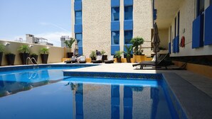 Une piscine sur le toit, agrémentée de chaises longues et de plantes en pot, entourée d’un bâtiment aux touches de bleu et doté d’un mur à motifs.