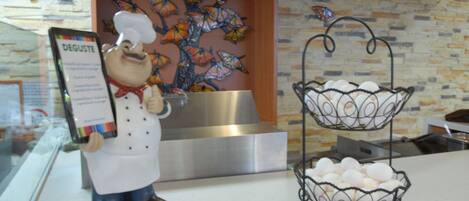 Una cocina con una figura decorativa de un chef sosteniendo un cartel, una cesta de metal con huevos y un cuadro de una mariposa en la pared.