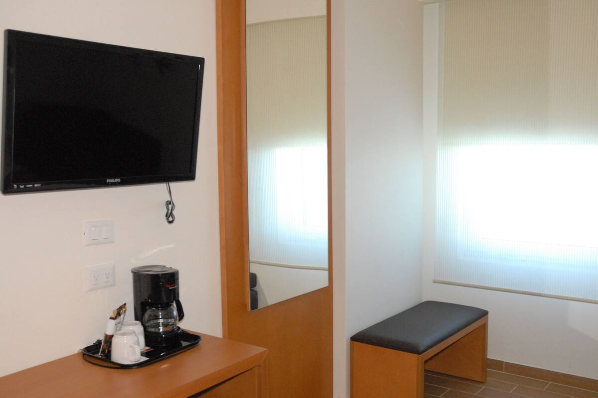 Habitación de hotel con televisión de pantalla plana, cafetera, una mesita, un banco y una ventana con persianas.