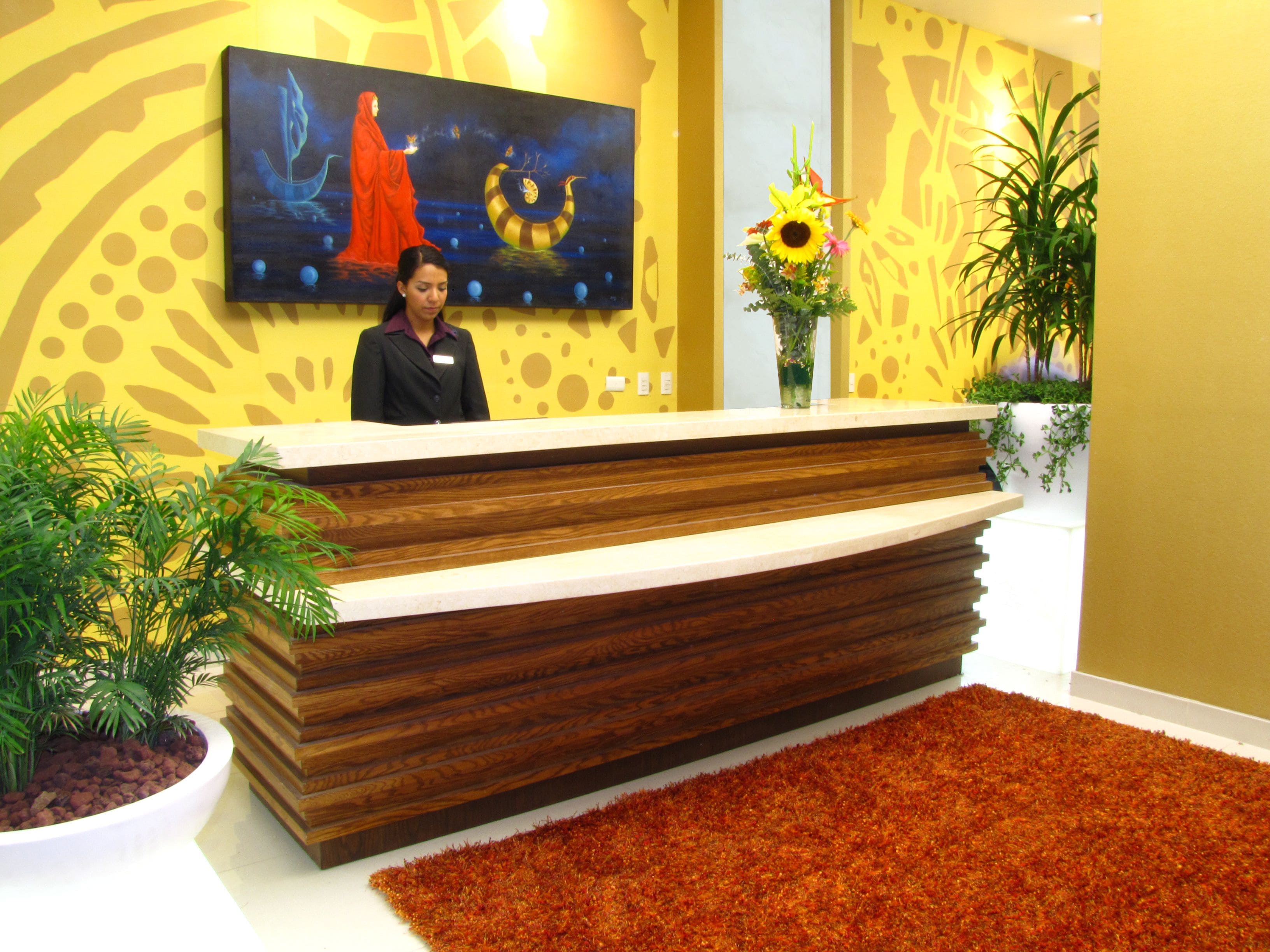 A reception desk with a wooden finish, a receptionist, a painting on the wall, and a vase with flowers.