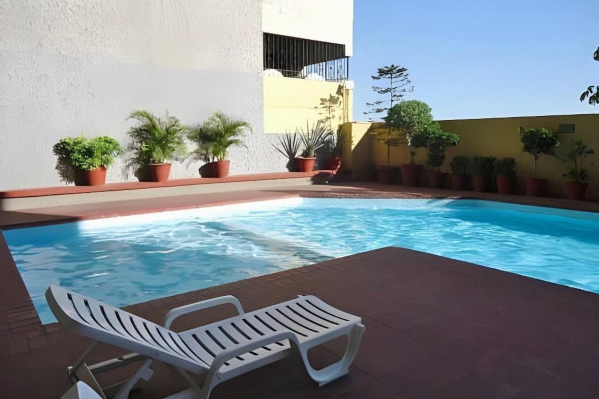 Une piscine entourée de chaises longues, de plantes en pot et d’un bâtiment.