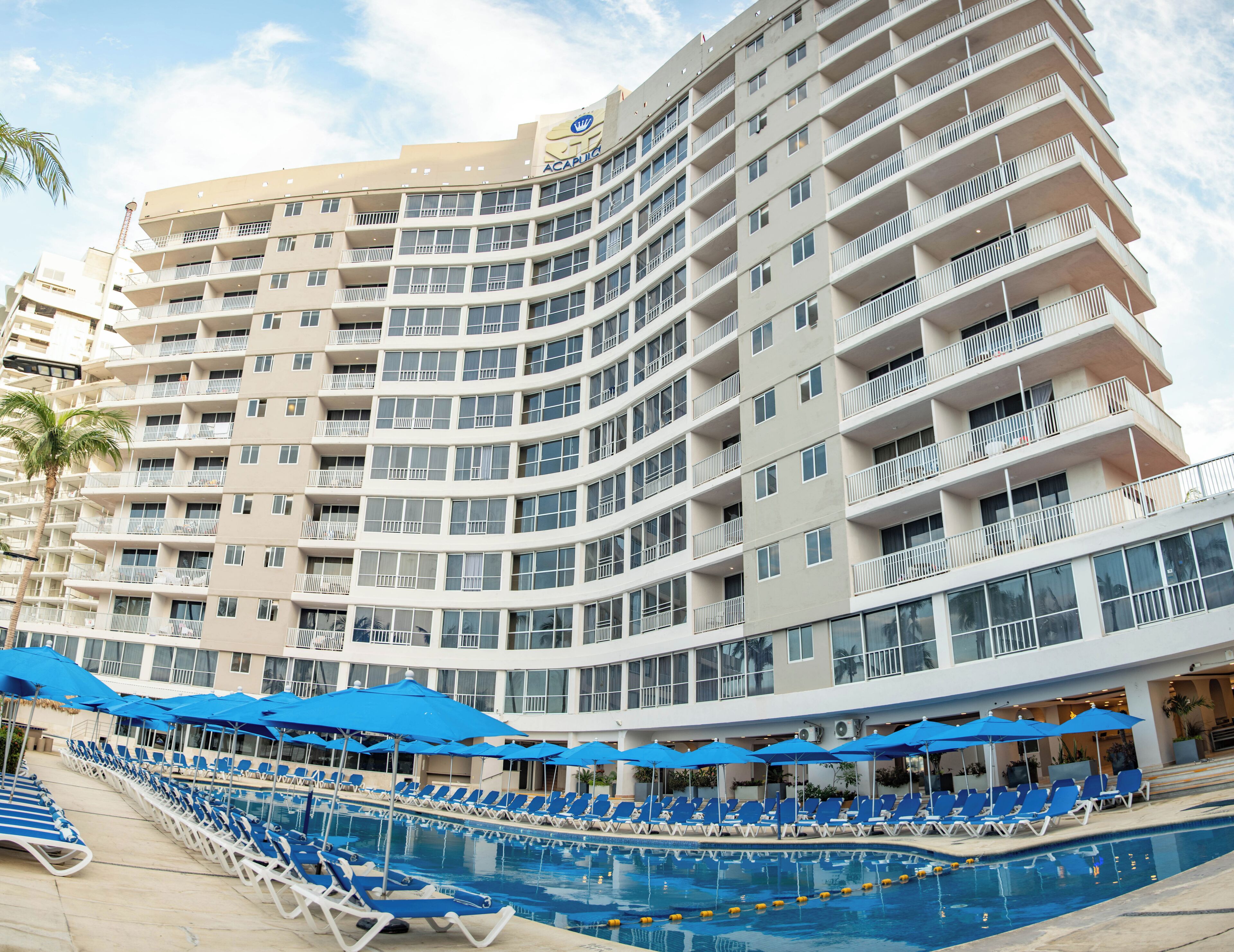 A multi-story hotel with a pool area featuring blue umbrellas and lounge chairs.