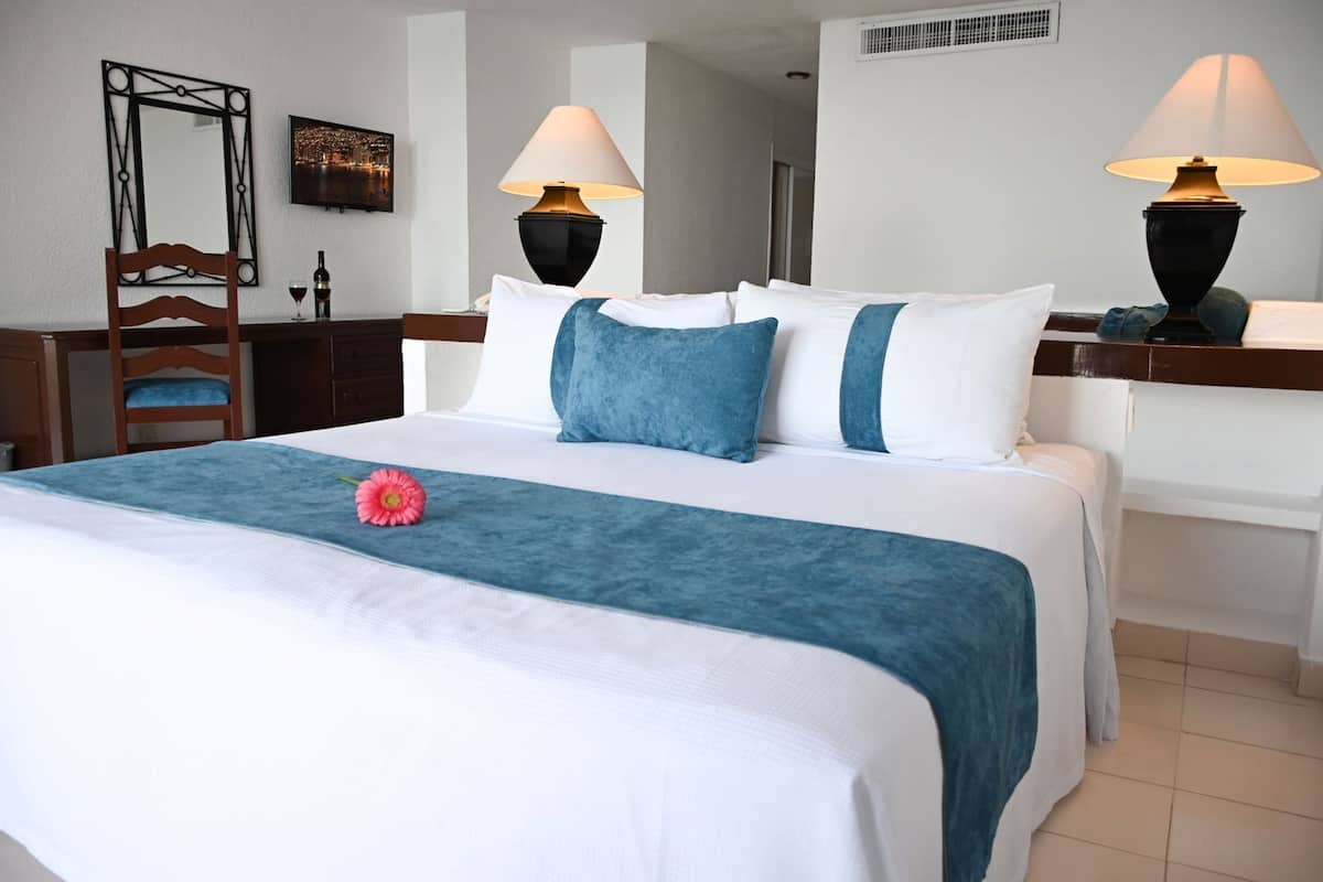 A hotel room with a bed, two bedside lamps, a wooden chair, a desk, and a mirror.