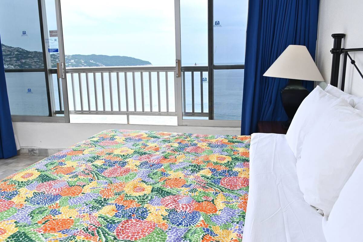 A hotel room with a large window, a bed with white bedding, and a view of the sea.