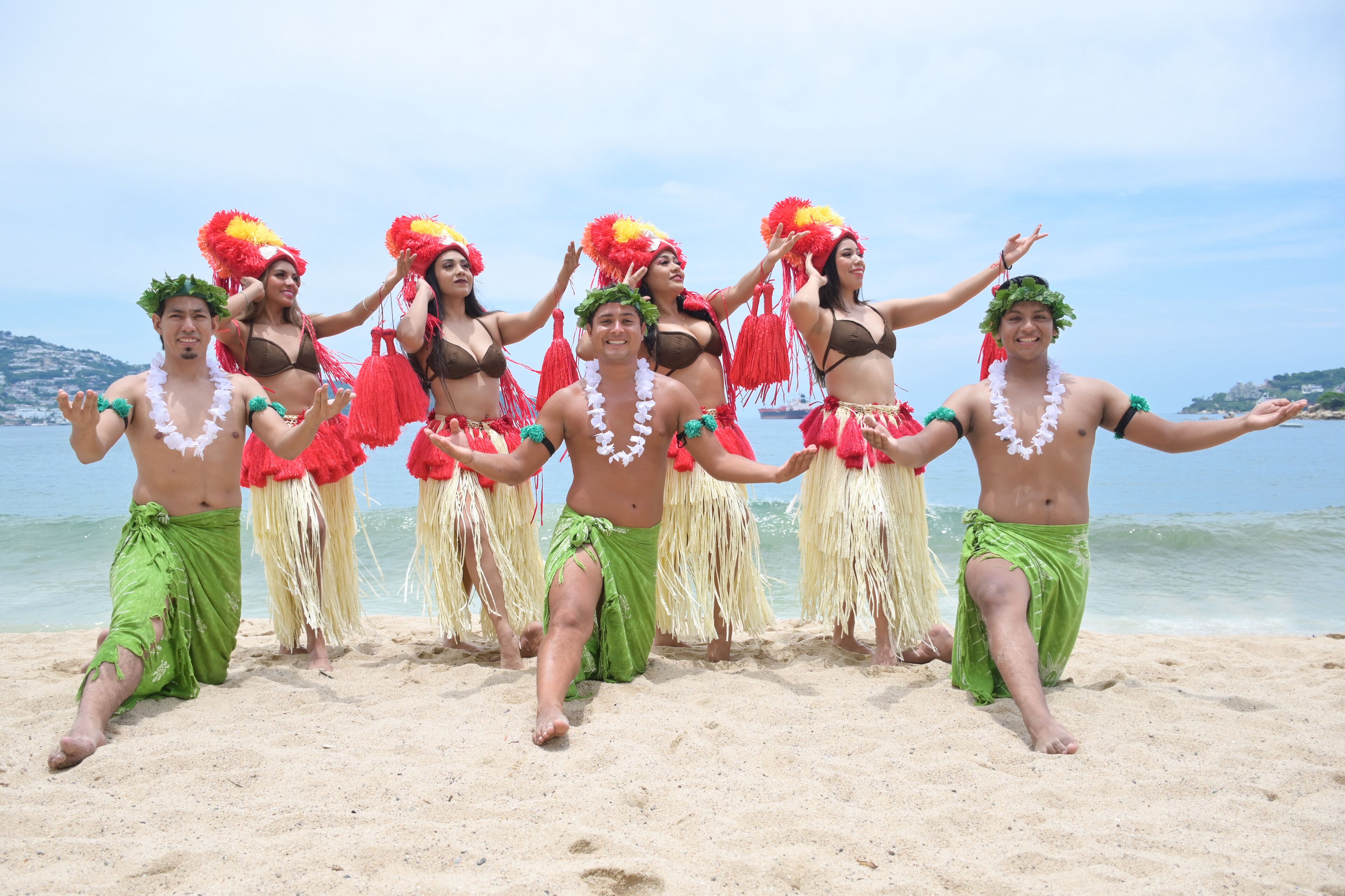 Eine Gruppe von Menschen tanzt am Strand, im Hintergrund ein Gewässer und eine Küstenlinie.