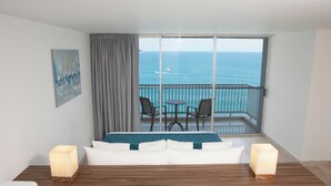 Una habitación de hotel con balcón, una cama con ropa blanca, una mesa de madera, dos sillas y un cuadro en la pared.