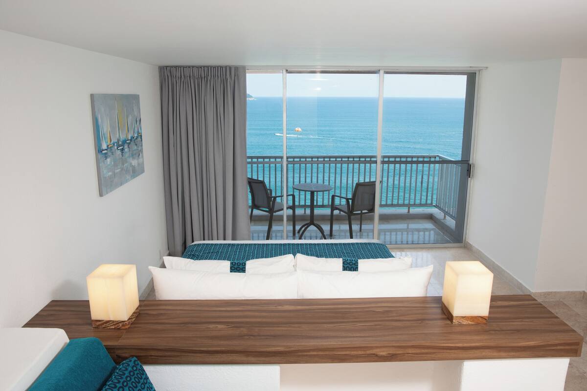 A hotel room with a balcony, a bed with white linens, a wooden table, two chairs, and a painting on the wall.