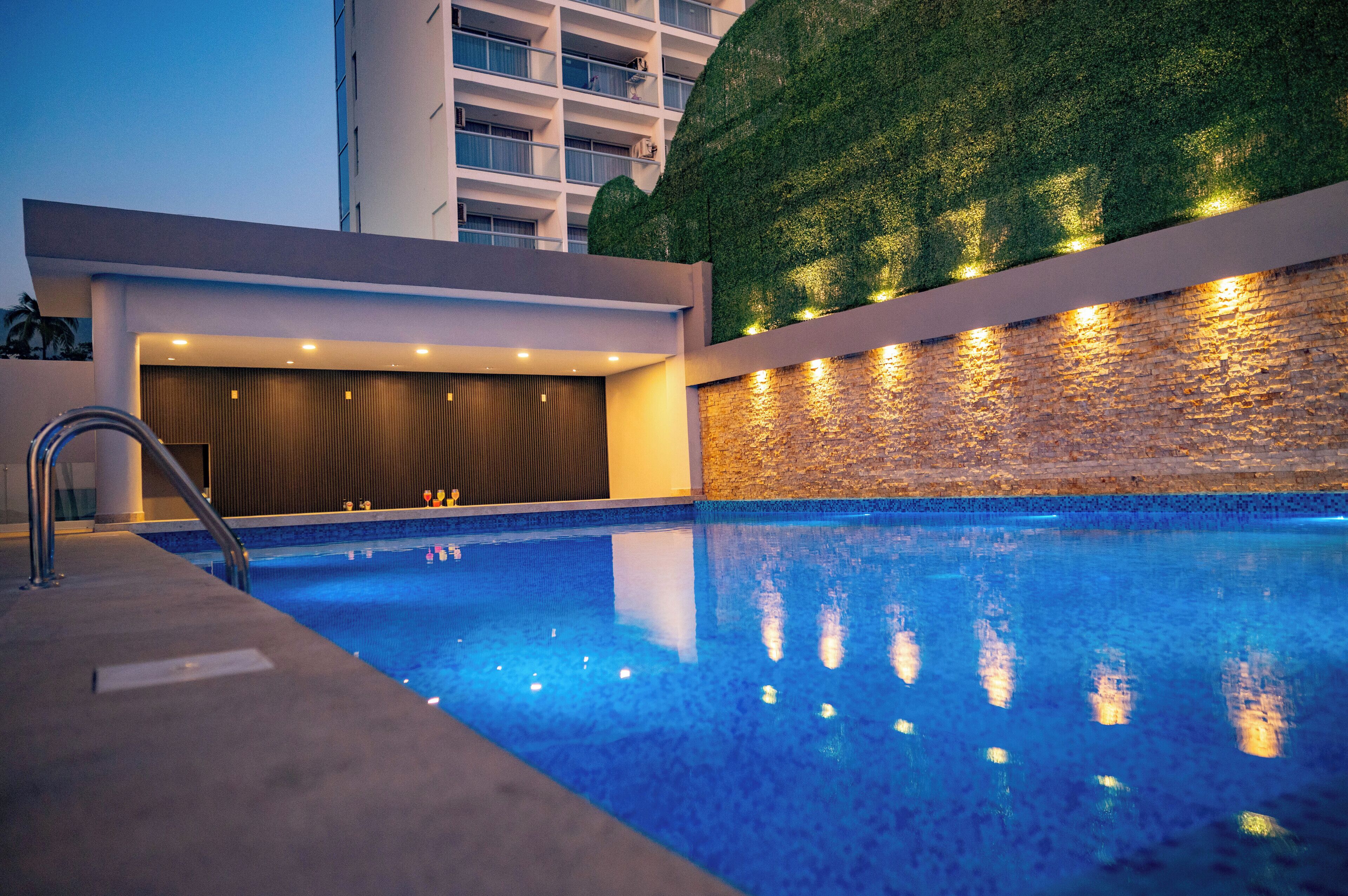 En swimmingpool med en mur av sten och en grön vägg, omgiven av en modern byggnad med en balkong.