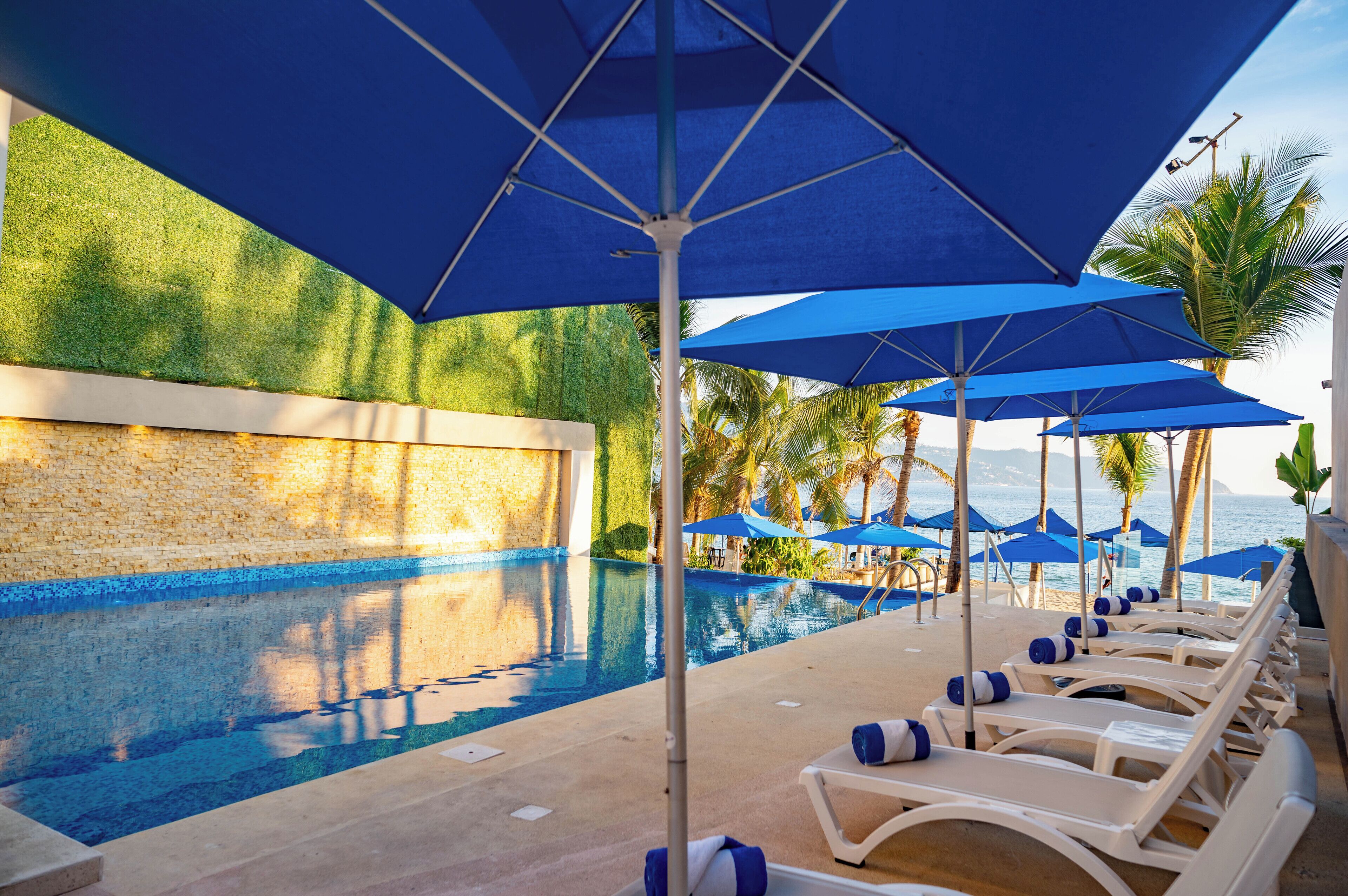 Uma área à beira da piscina com espreguiçadeiras e guarda-sóis azuis, uma piscina límpida e vista para o mar.