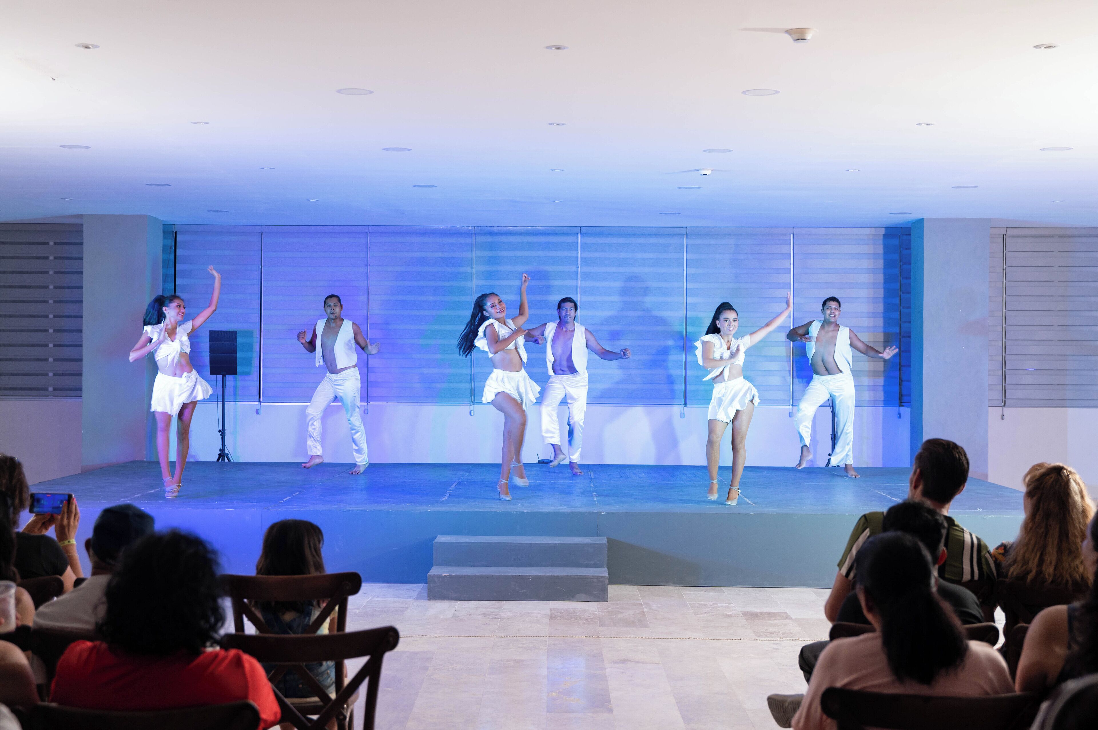 A stage with performers in white outfits, an audience seated in front, and a modern interior with a blue-lit background.