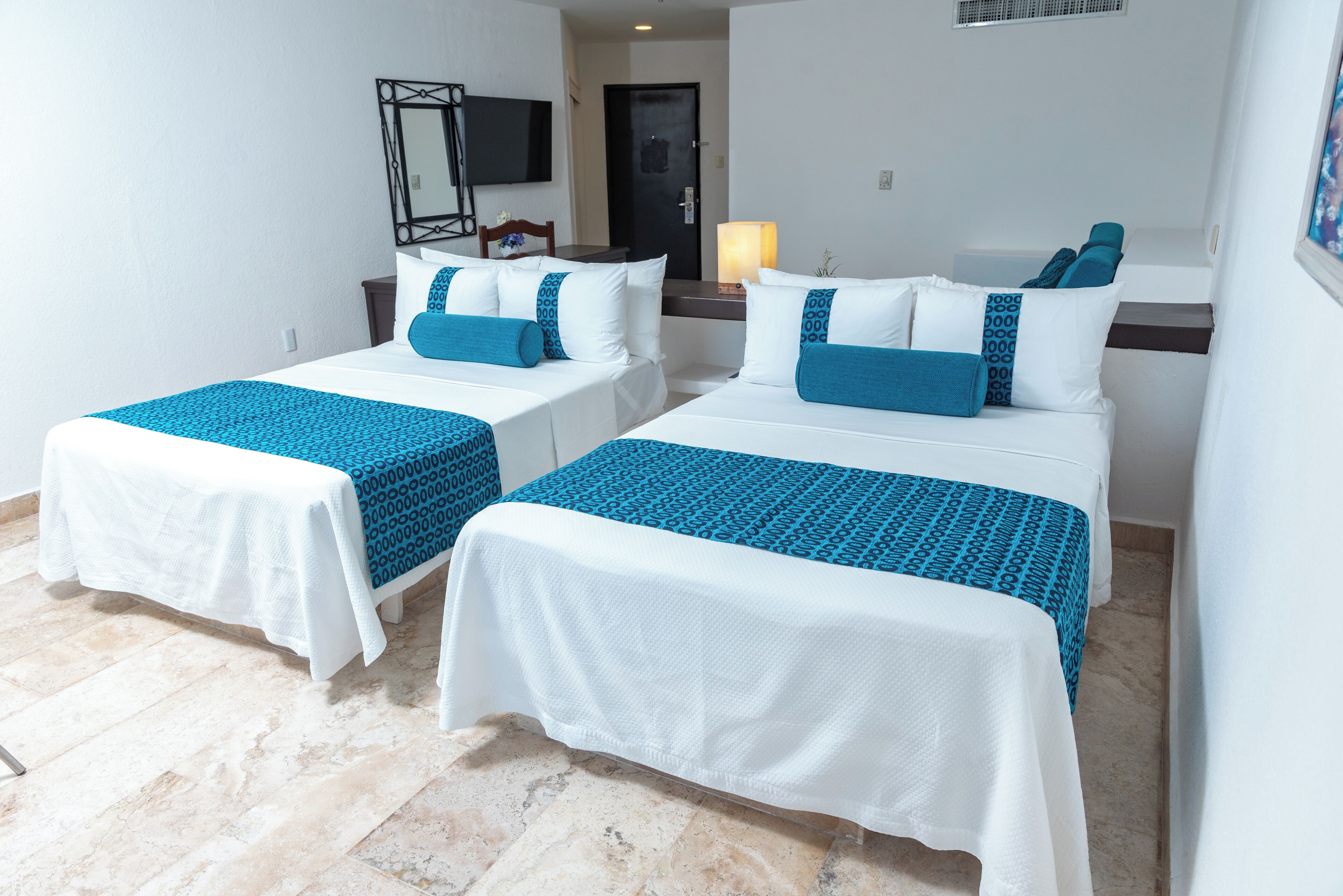 Habitación de hotel con dos camas individuales, cada una con colcha blanca y azul estampada, una mesita de noche con lámpara y un televisor montado en la pared.