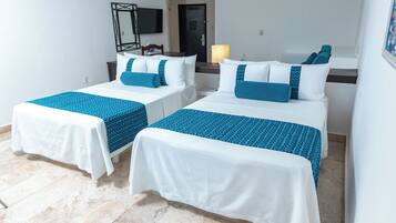 Habitación de hotel con dos camas individuales, cada una con colcha blanca y azul estampada, una mesita de noche con lámpara y un televisor montado en la pared.