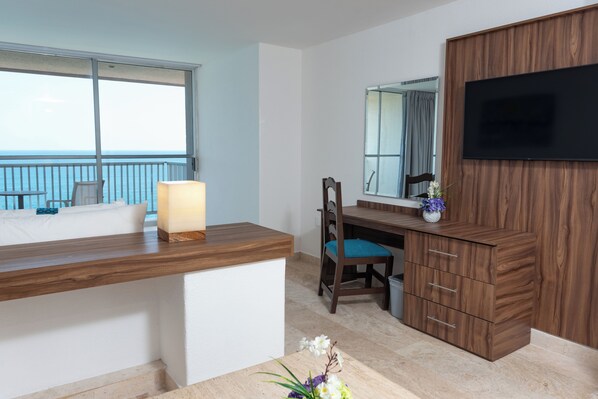 Habitación de hotel con un escritorio de madera, una silla, un televisor montado en un panel de madera, un ventanal con vistas al mar y una mesita con un jarrón de flores.