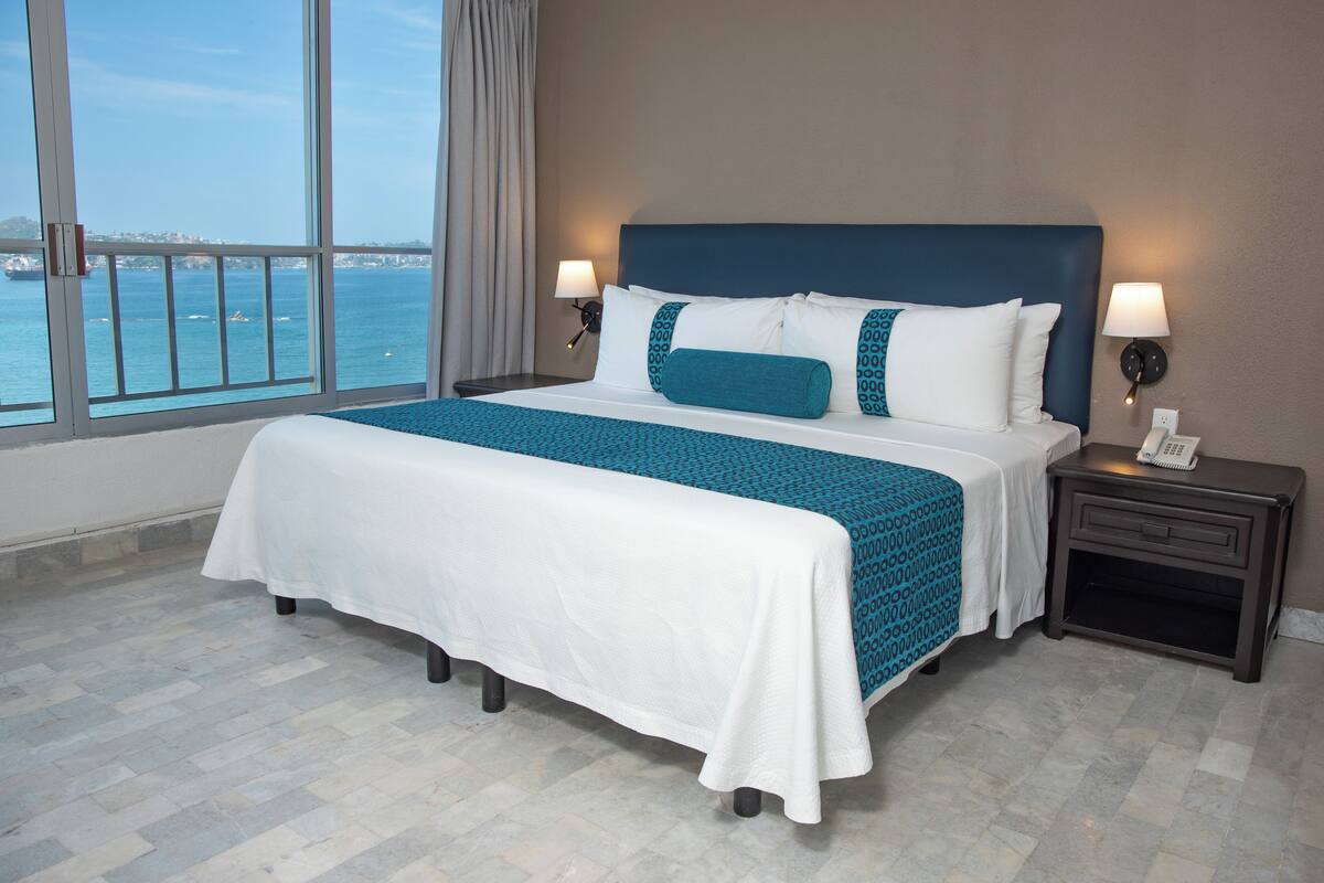 A hotel room with a large bed, two bedside tables, a view of the sea, and a cityscape in the distance.