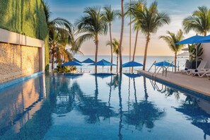 A swimming pool with clear blue water, surrounded by palm trees and blue umbrellas.