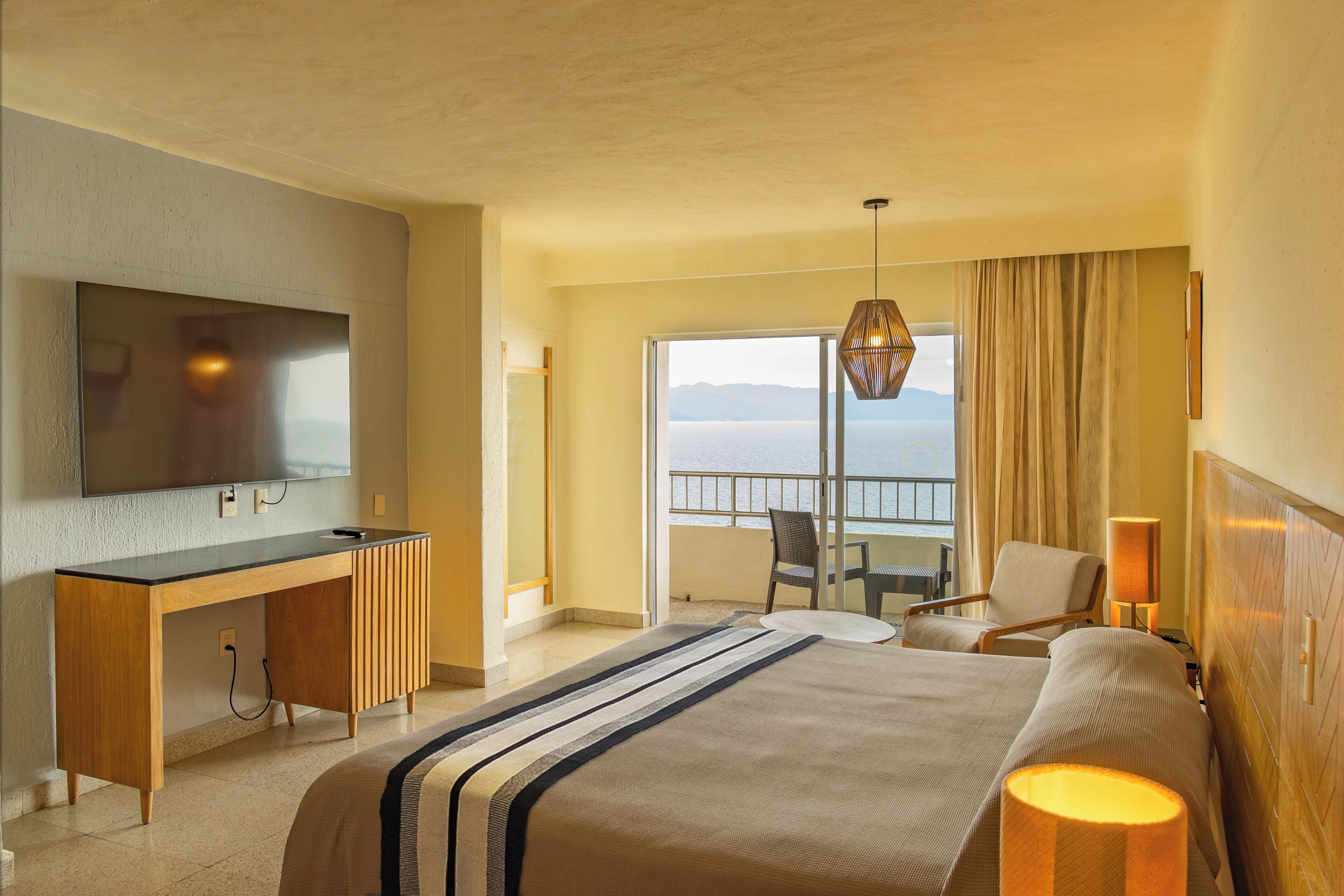 Habitación de hotel con cama, escritorio, televisión, balcón con vistas y una silla.