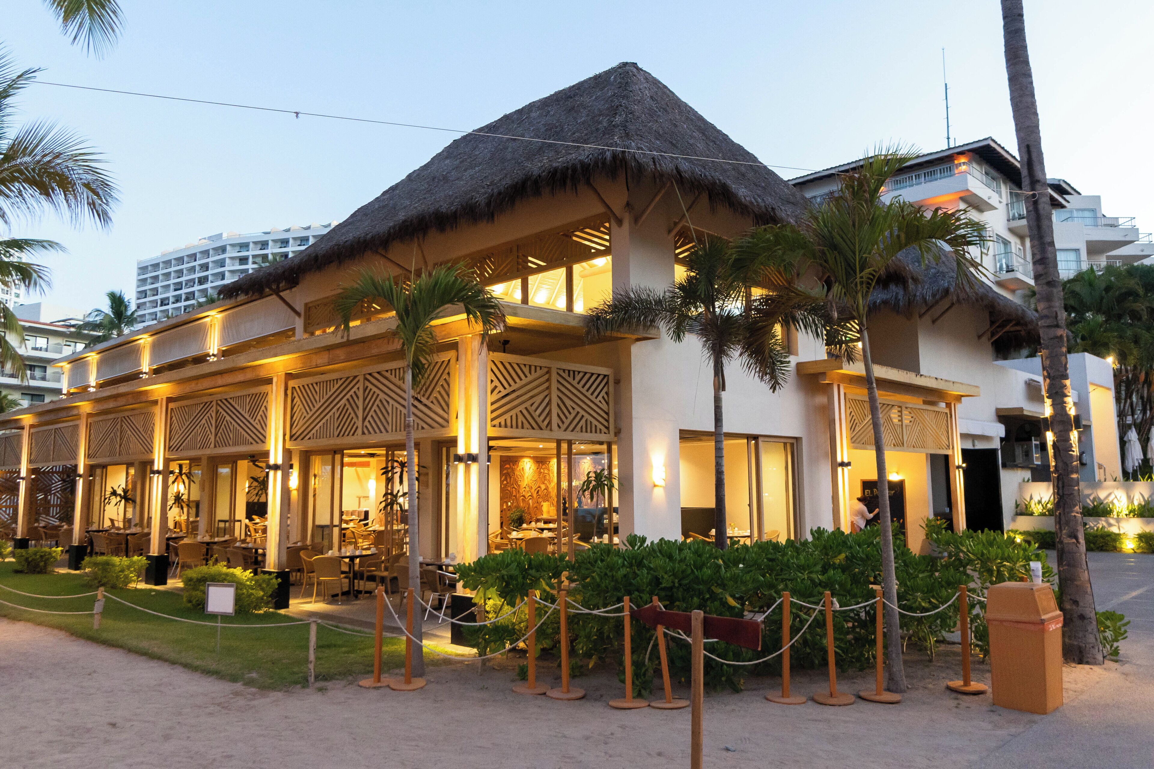 A hotel with a thatched roof, outdoor seating, and palm trees.