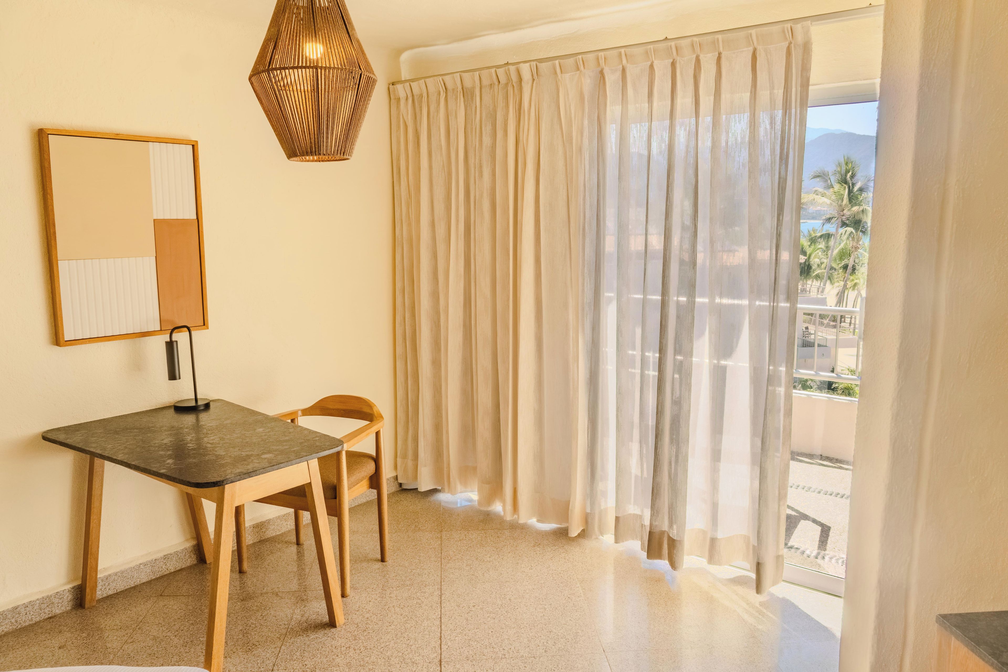 Ein Zimmer mit Schreibtisch, Stuhl und einem großen Fenster mit transparenten Vorhängen.