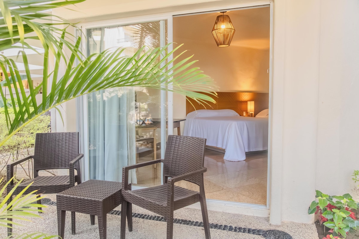 A hotel room with a bed, a nightstand, and a lamp. There is a sliding glass door leading to a balcony with two wicker chairs and a potted plant.