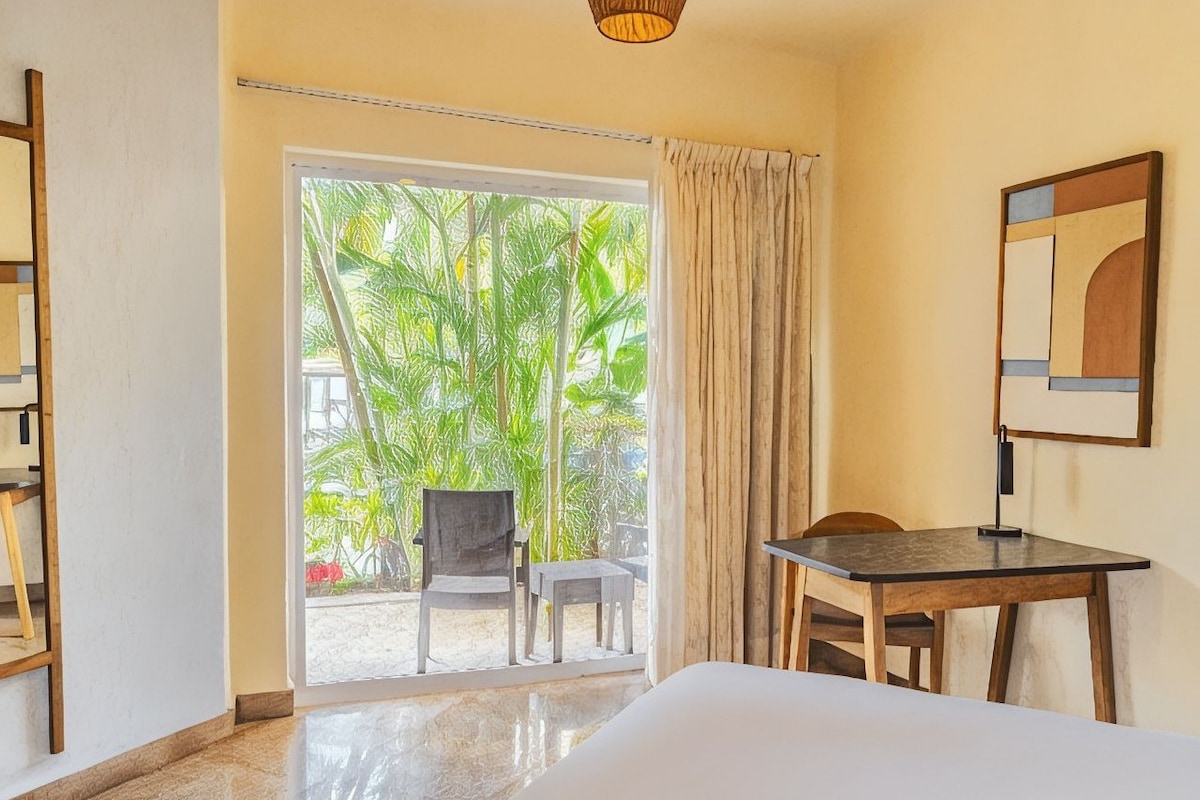 A hotel room with a bed, a desk, a chair, and a view of greenery outside.