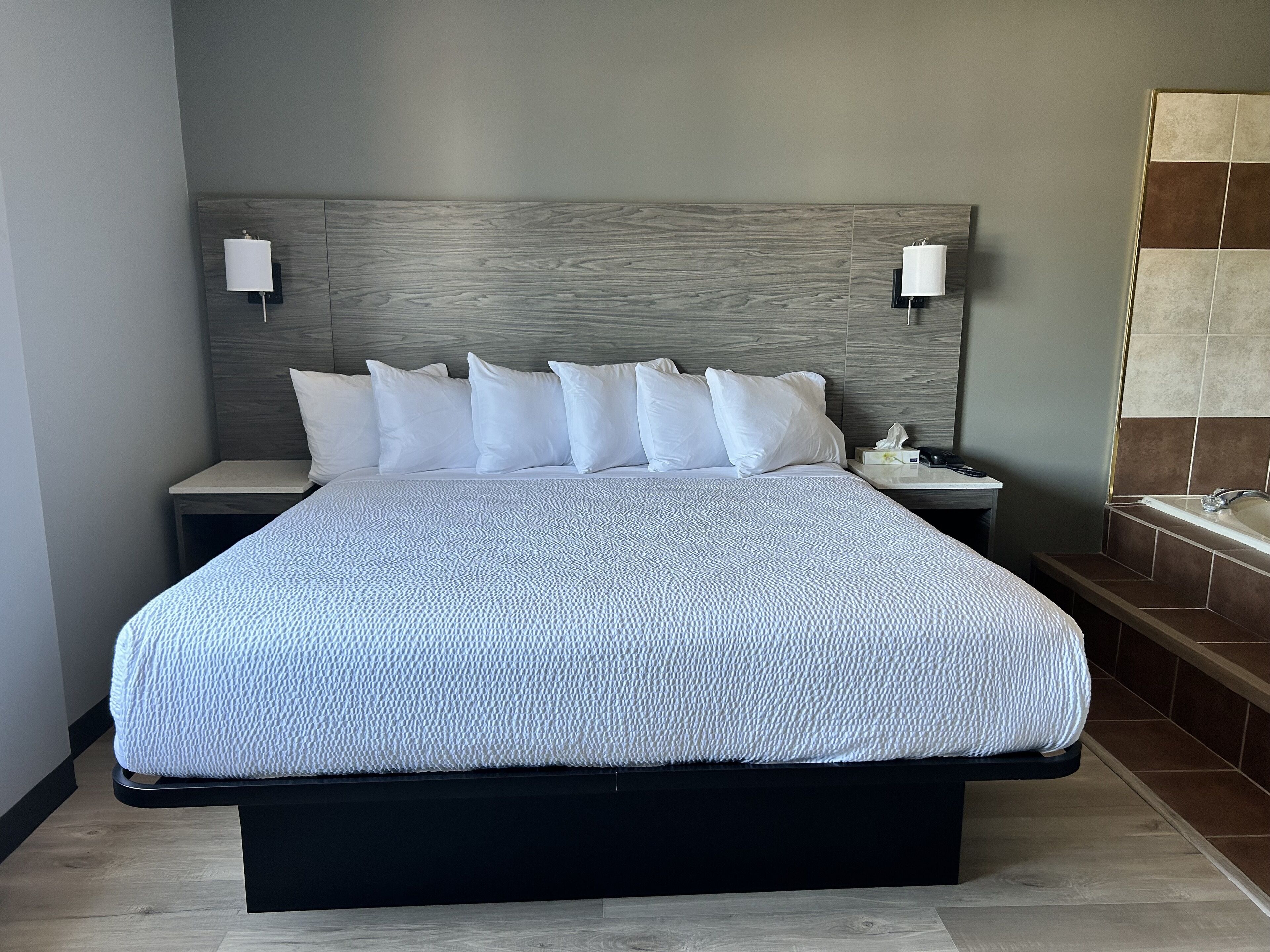 Un lit bien fait, recouvert d’une housse blanche à texture intéressante, deux tables de chevet avec des lampes, et une salle de bain dotée d’une baignoire et de murs carrelés.