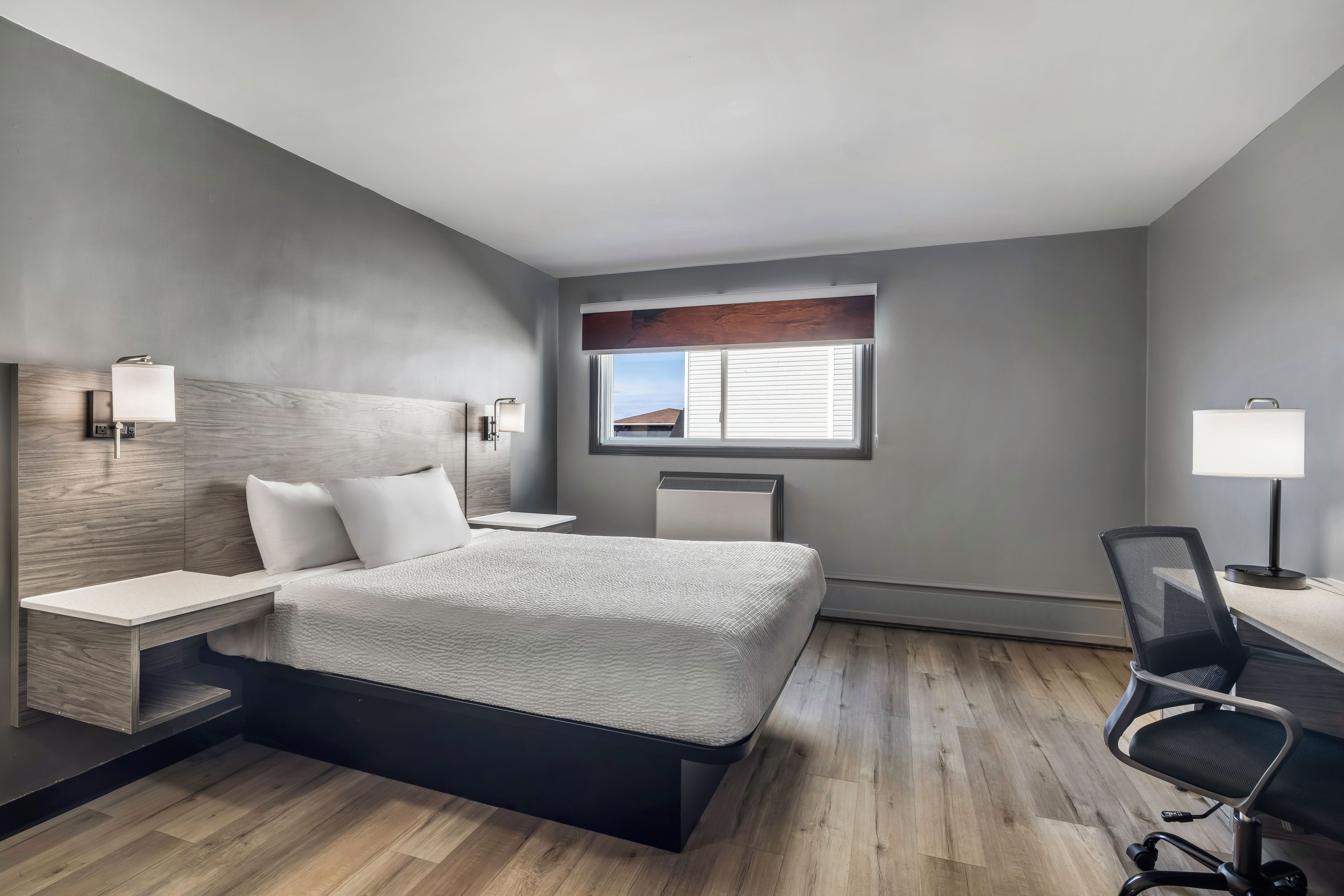 A modern hotel room with a bed, bedside tables, a desk, a chair, and a window with blinds.
