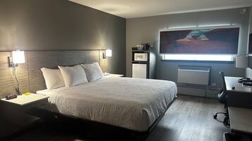 A modern hotel room with a large bed, bedside table, lamp, and a desk with a chair.
