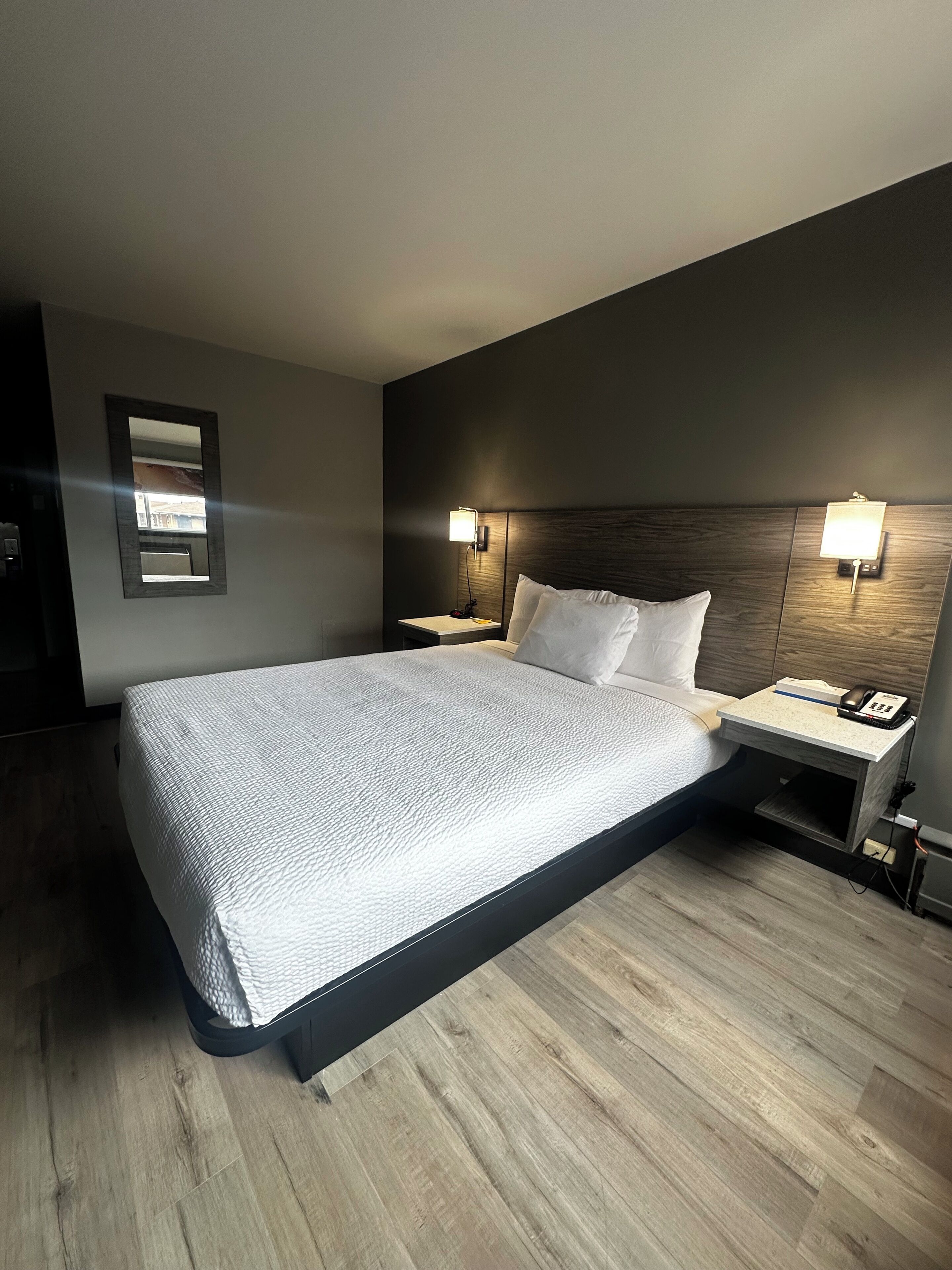 Une chambre d’hôtel moderne avec un grand lit, deux tables de chevet avec des lampes, une tête de lit en bois et un miroir au mur.