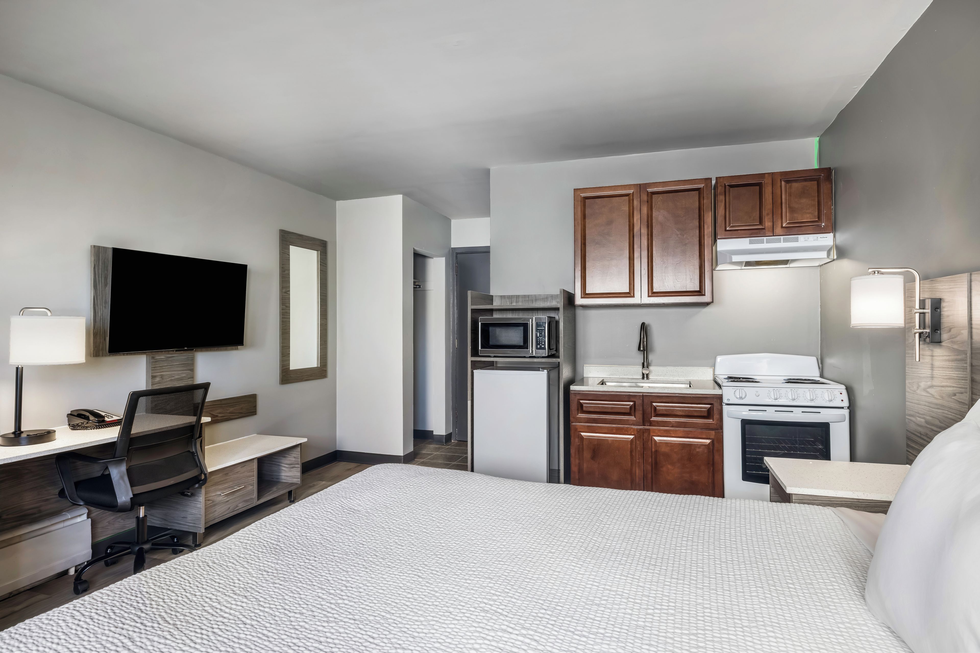 A hotel room with a bed, desk, chair, television, kitchenette, and a bathroom.