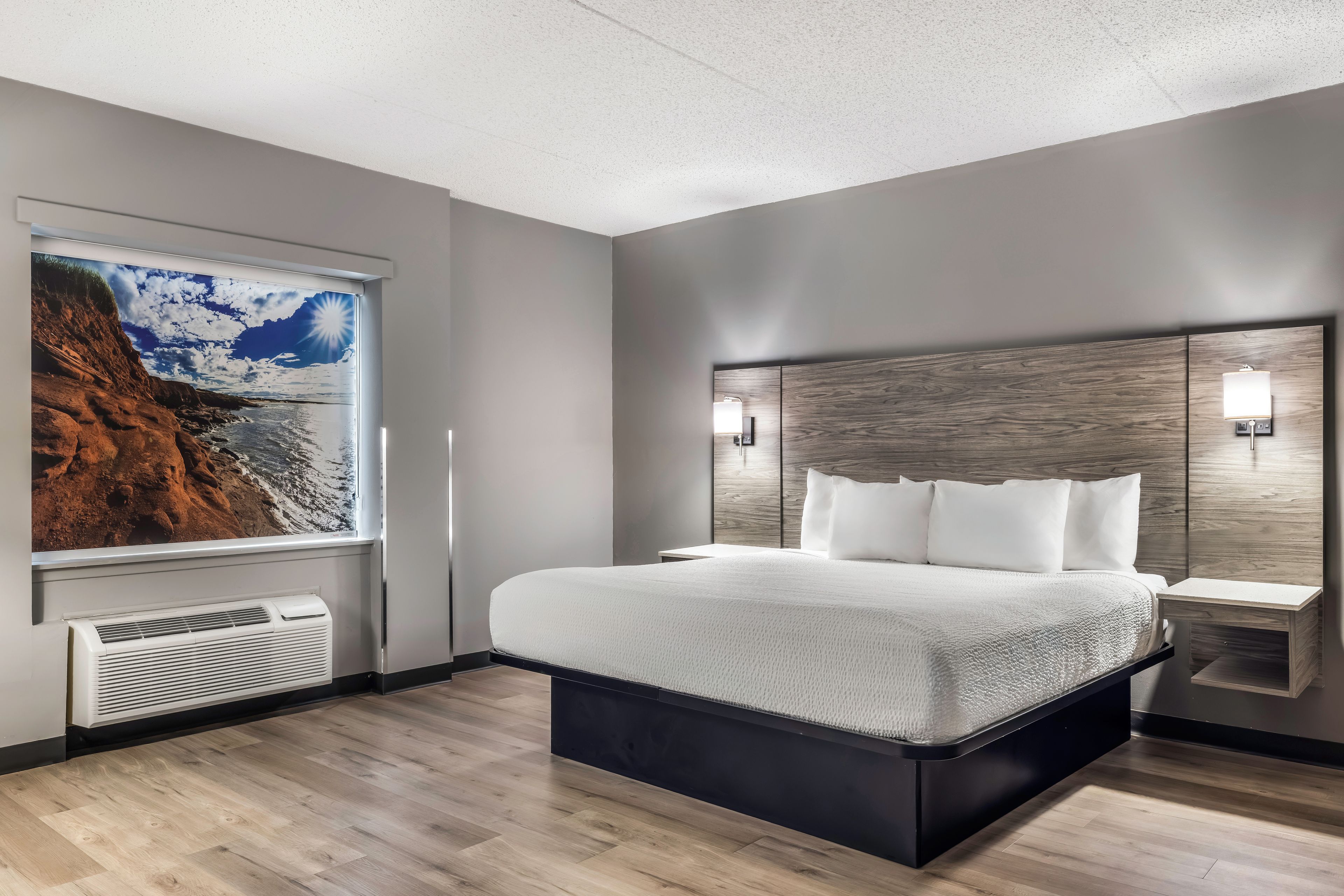 A modern hotel room with a large bed, a wall-mounted air conditioner, a window with a scenic view, and a nightstand with a lamp.